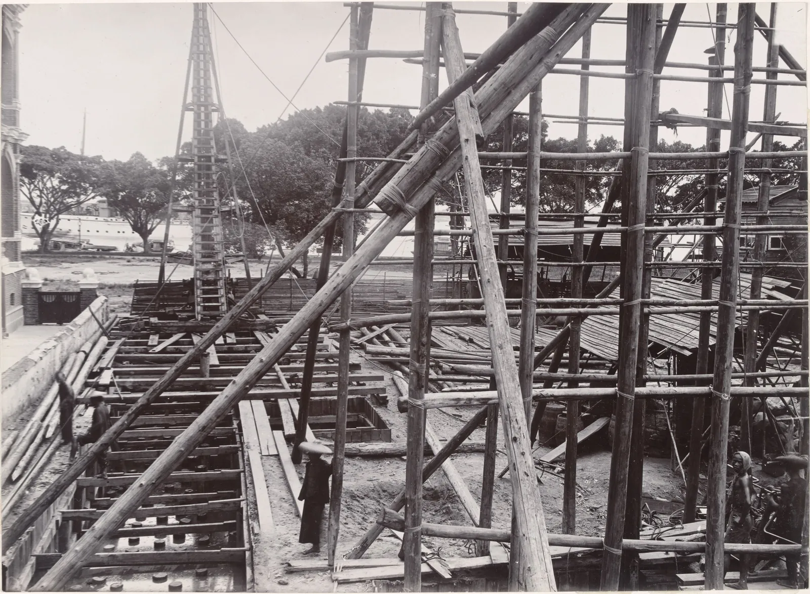 Bamboo scaffolding and timber beams over a building site, with a workman by a ladder.