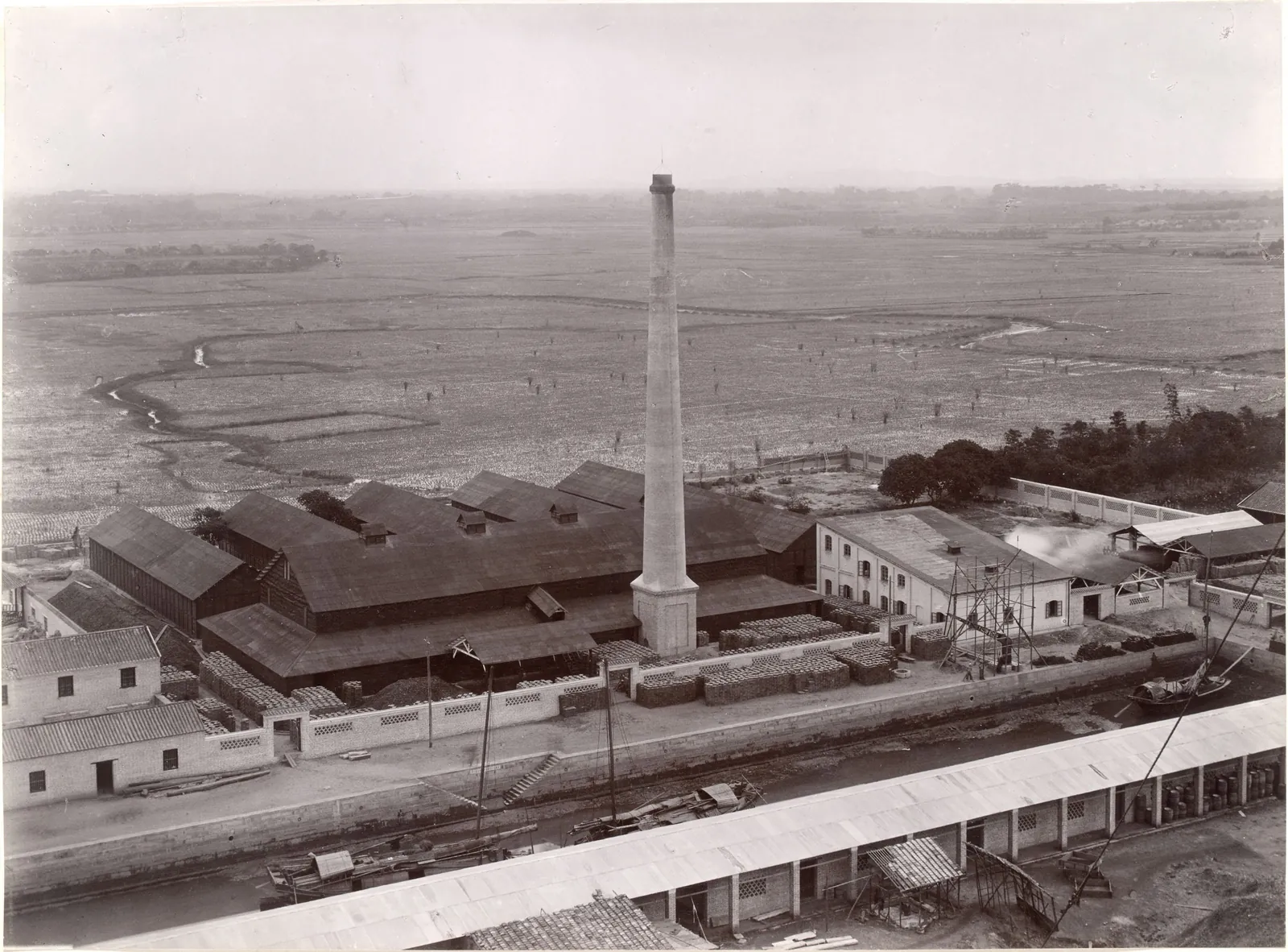 A factory with a tall chimney and long sheds beside the river, seen from above, flat country behind.