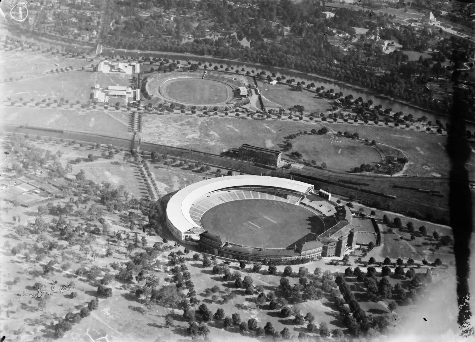 The Melbourne Cricket Ground from the air, a long curved stand with a white roof on its far side, and trees all round.