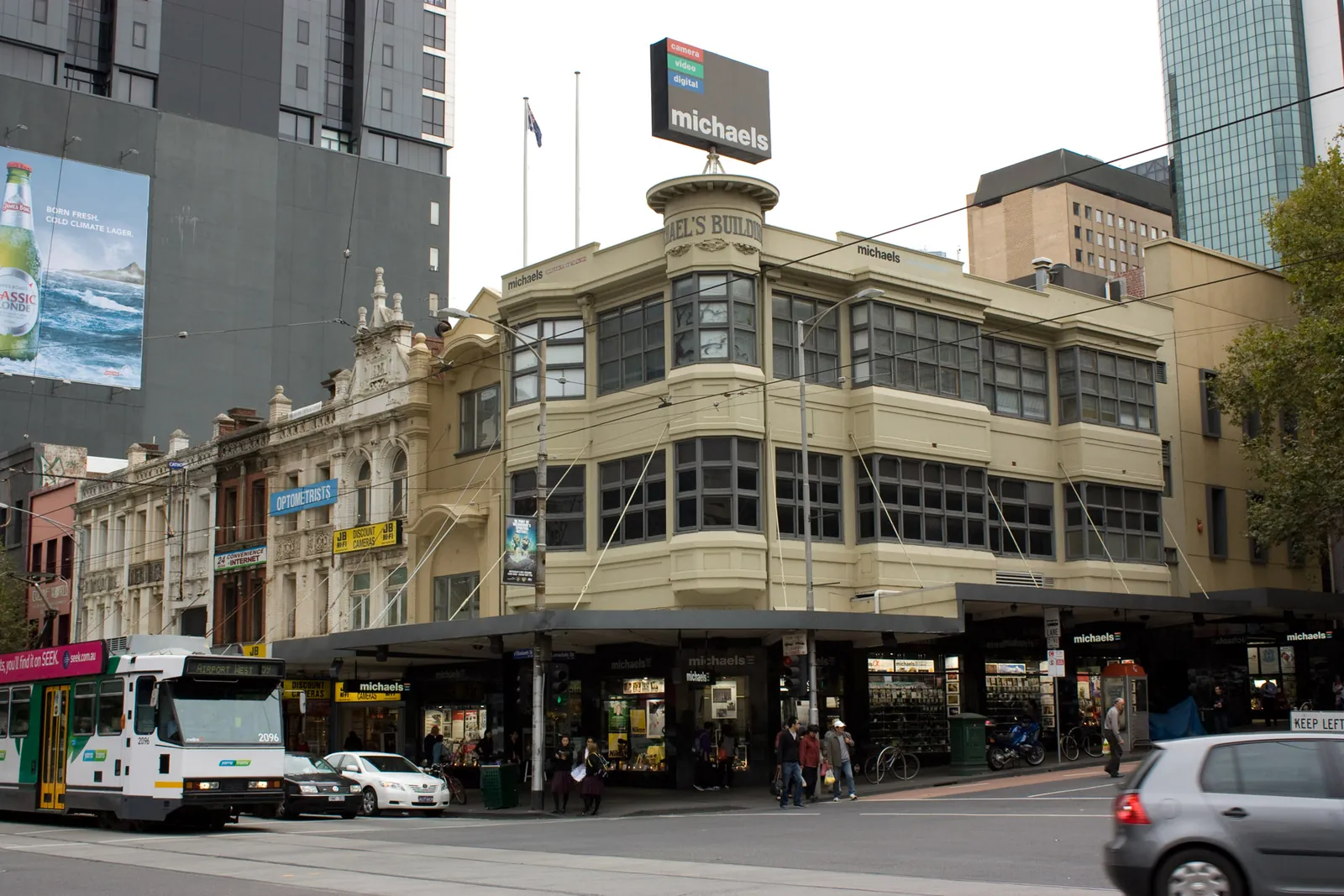 A cream corner building with bay windows and a corner tower, with a tram passing in front.