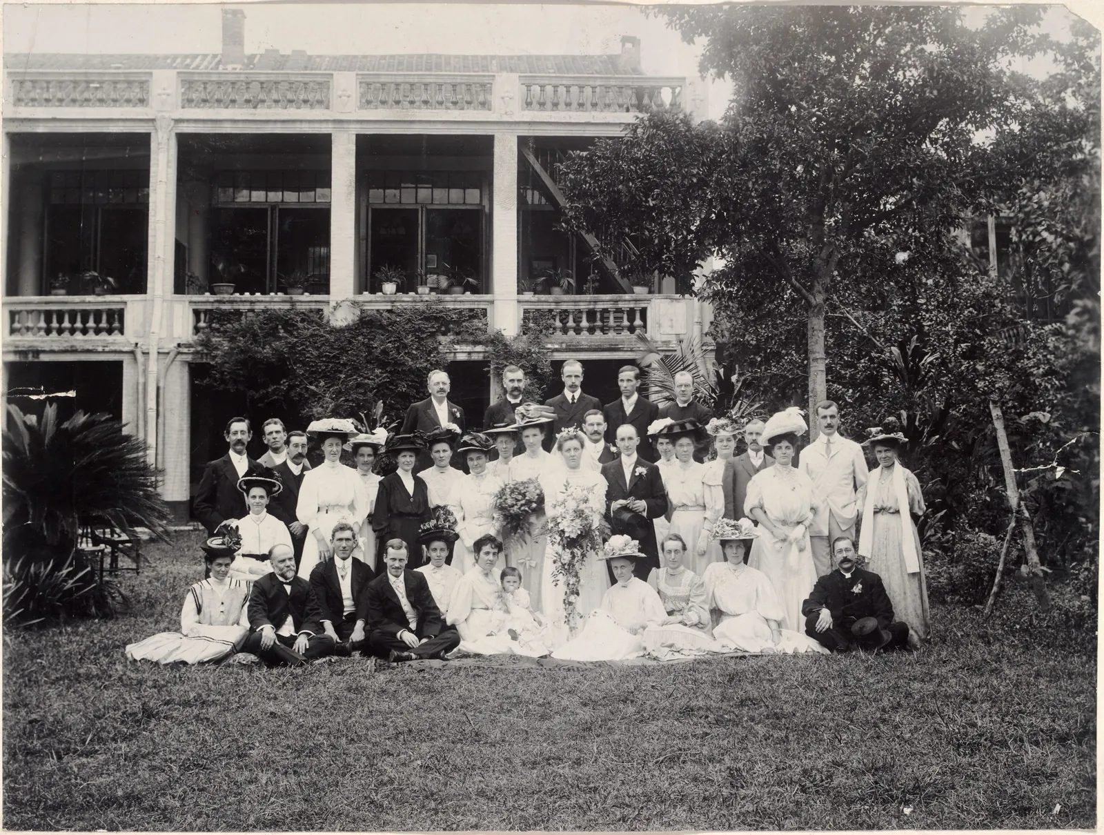 A large wedding party on a lawn in front of a two-storey verandahed building, the women in white dresses and hats.