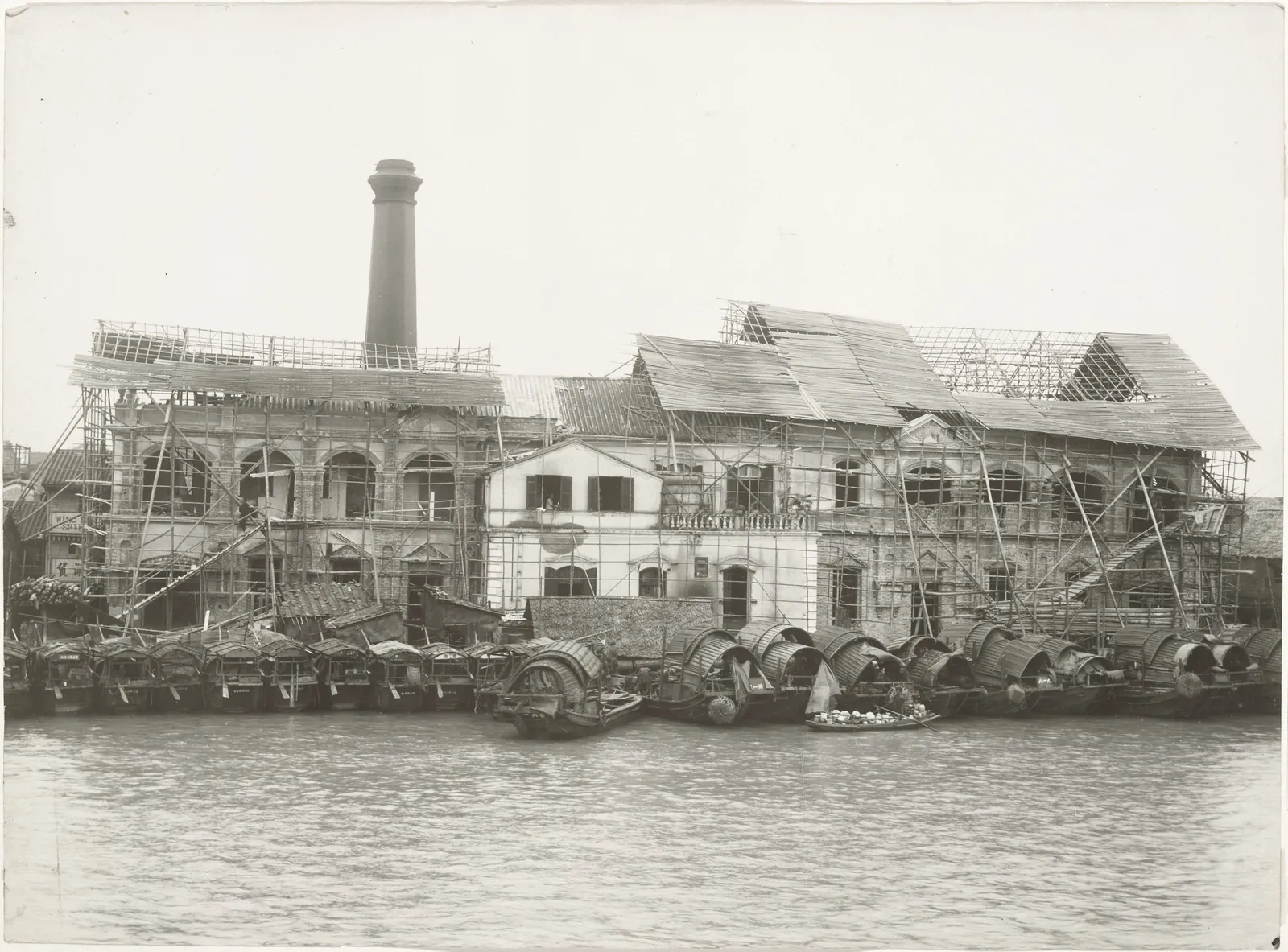 The power station being built, its roof still under mat sheds, with sampans moored in front.
