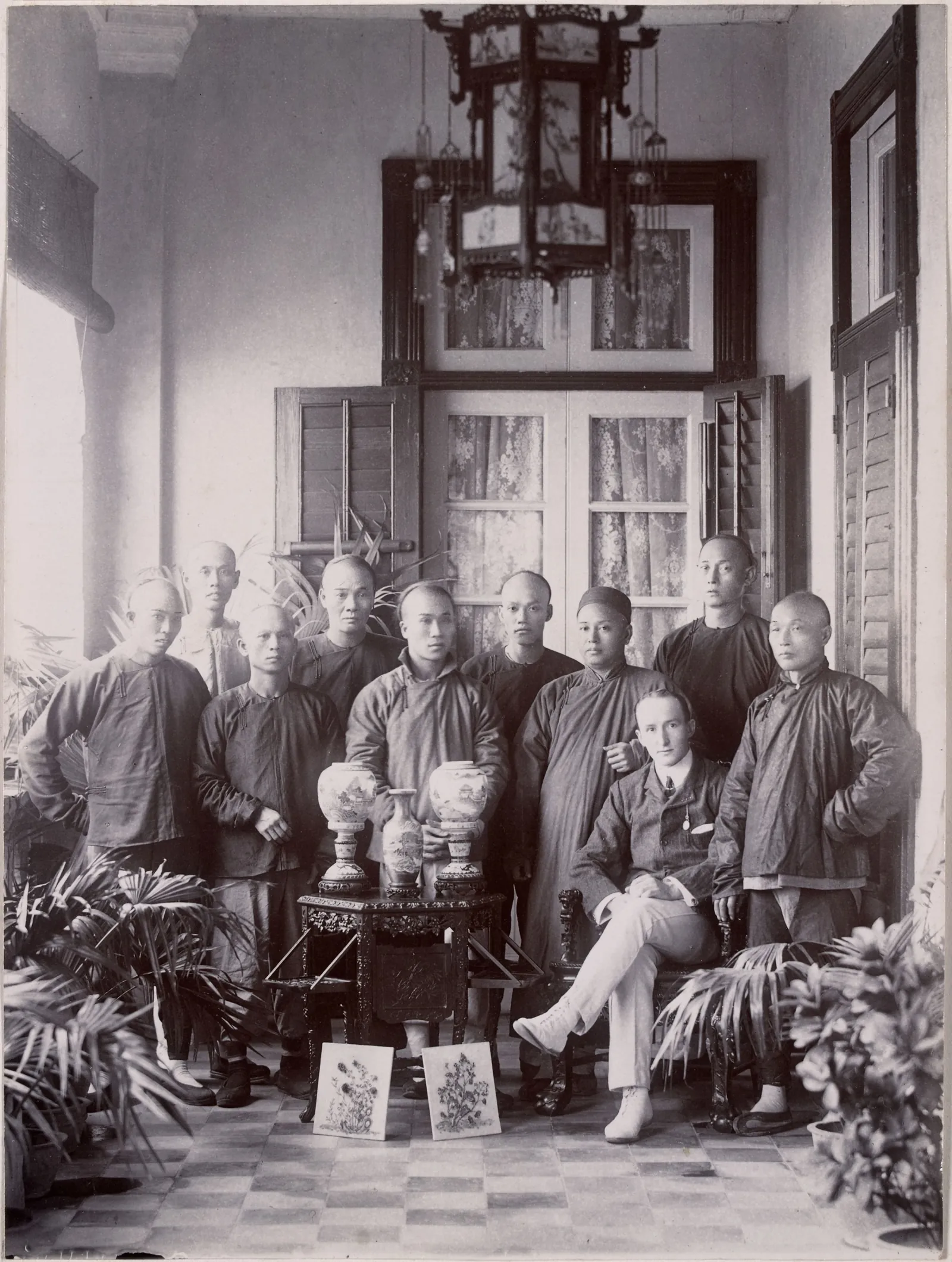About fifteen men in Chinese jackets and caps grouped round two seated Europeans, under a hanging lantern, with porcelain vases on a table.