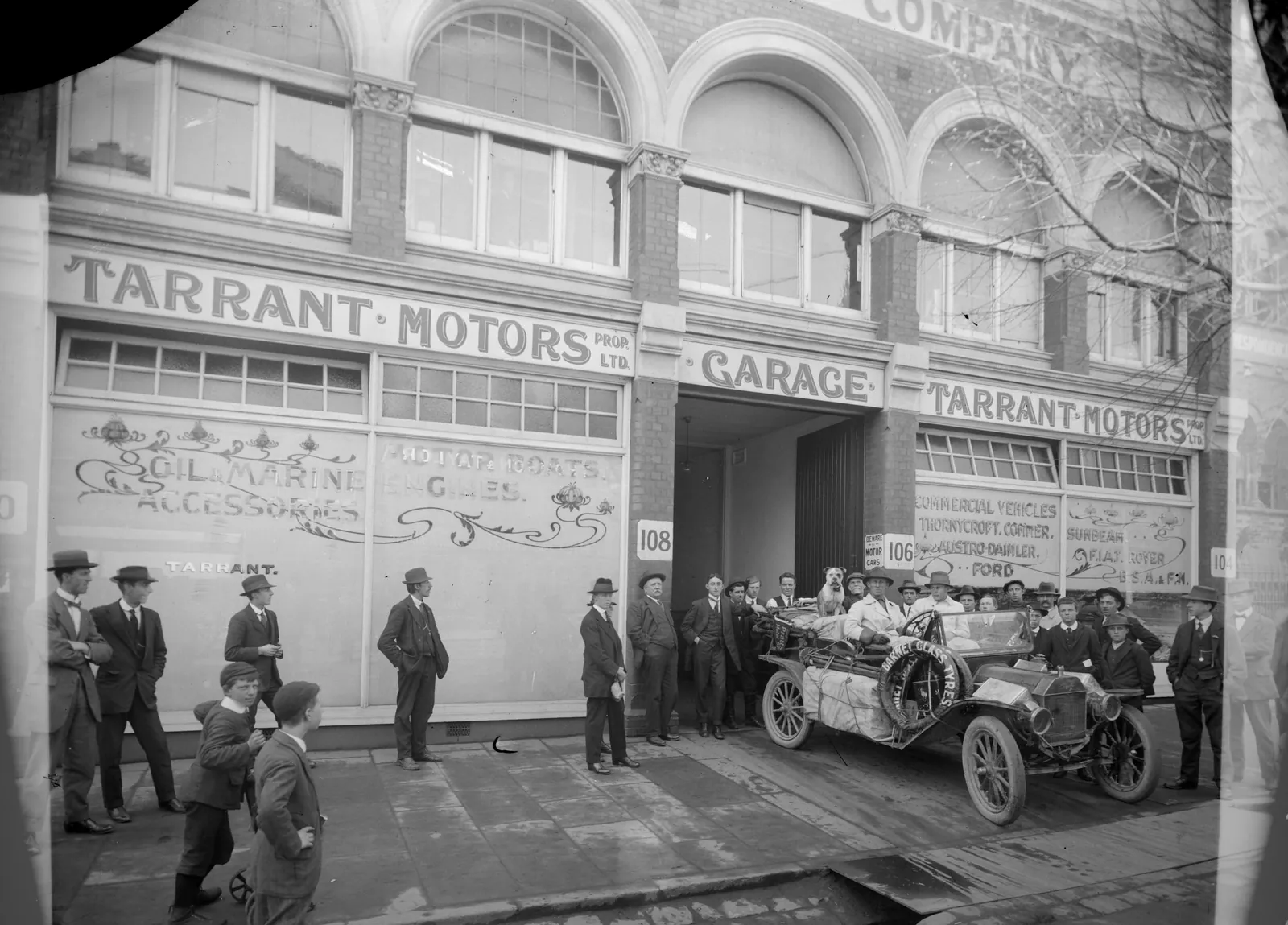 A brick garage front lettered Tarrant Motors, with an open car piled with luggage and a bulldog in the entrance, and men and boys in hats on the footpath.